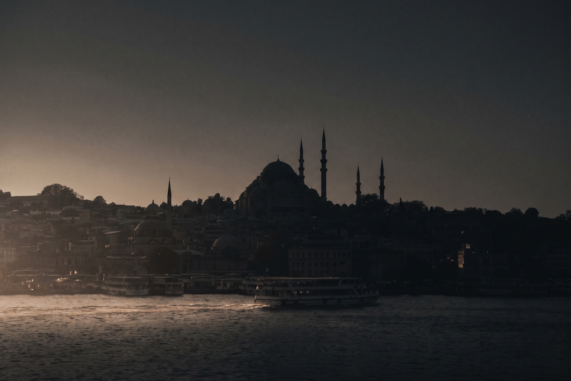 Istanbul skyline at dusk from the water, with domes and minarets in silhouette above a ferry wake.