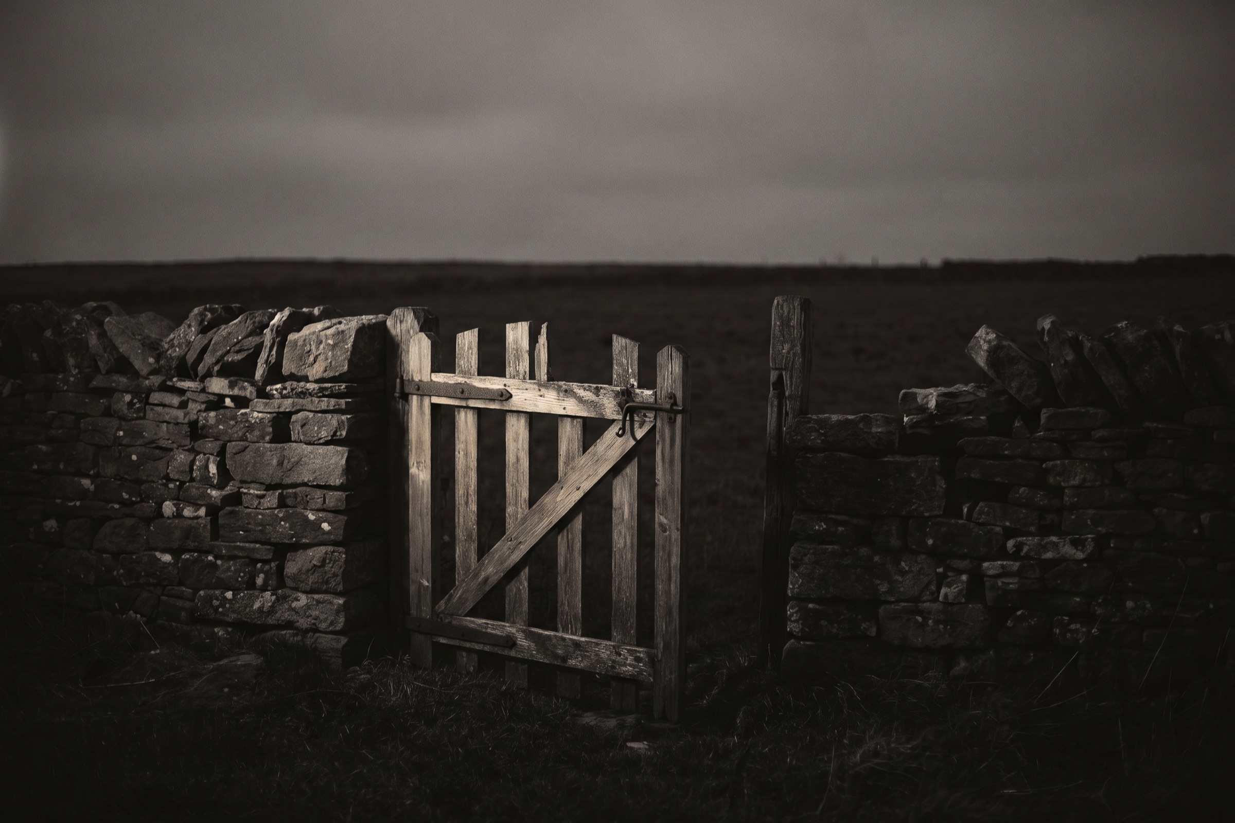 Plain wooden gate standing half open in a dry stone wall, with an empty field beyond under a gray sky.